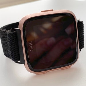 May include: A rose gold Fitbit smartwatch with a black woven band. The watch face is black and displays the Fitbit logo.