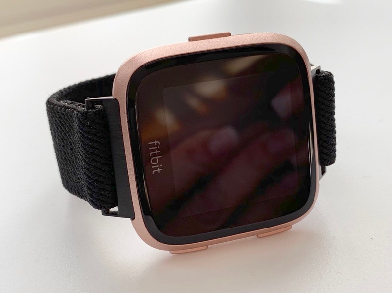 May include: A rose gold Fitbit smartwatch with a black woven band. The watch face is black and displays the Fitbit logo.