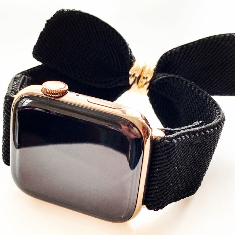 May include: A rose gold Apple Watch with a black elastic band that has a bow on the top.