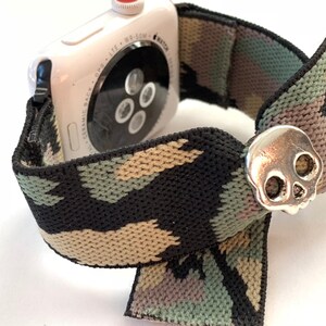 May include: A white Apple Watch with a black, green, and tan camouflage patterned elastic band. The band has a silver skull charm.