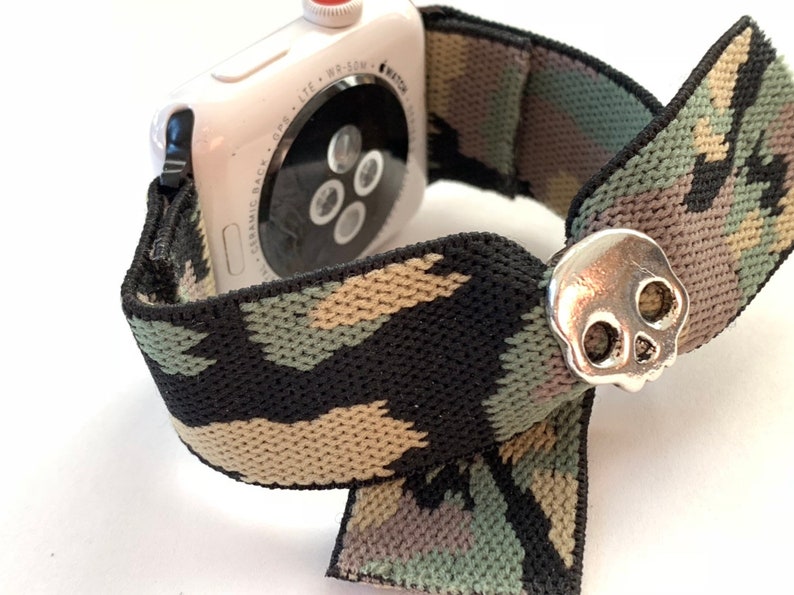 May include: A white Apple Watch with a black, green, and tan camouflage patterned elastic band. The band has a silver skull charm.