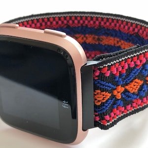 May include: A rose gold Fitbit smartwatch with a black and colorful patterned elastic band. The band has a geometric design with red, orange, blue, and white accents.