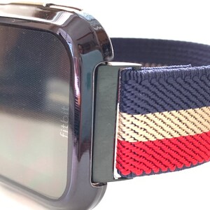 Puede incluir: Un reloj inteligente Fitbit negro con una banda el&aacute;stica a rayas azul marino, beige y rojo.