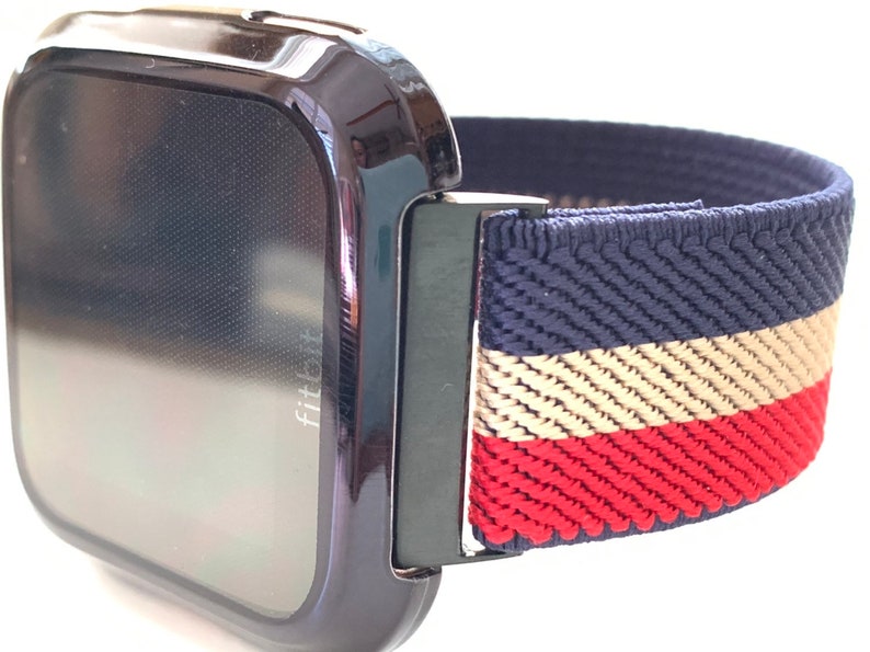 Puede incluir: Un reloj inteligente Fitbit negro con una banda el&aacute;stica a rayas azul marino, beige y rojo.