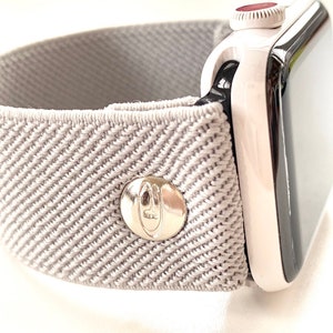 Puede incluir: Una banda elástica gris para Apple Watch con una hebilla plateada. La banda tiene un patrón texturizado y está diseñada para un look deportivo.