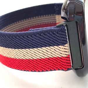 Puede incluir: Primer plano de una correa de reloj con un patr&oacute;n de rayas azul marino, beige y rojo. La correa est&aacute; hecha de un material el&aacute;stico y est&aacute; unida a un reloj negro.