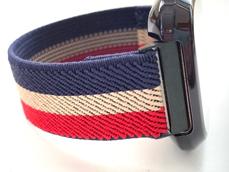 Puede incluir: Primer plano de una correa de reloj con un patr&oacute;n de rayas azul marino, beige y rojo. La correa est&aacute; hecha de un material el&aacute;stico y est&aacute; unida a un reloj negro.
