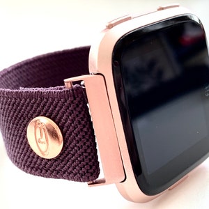 May include: A rose gold smartwatch with a dark gray screen and a dark purple elastic band. The band has a gold button with a logo on it.