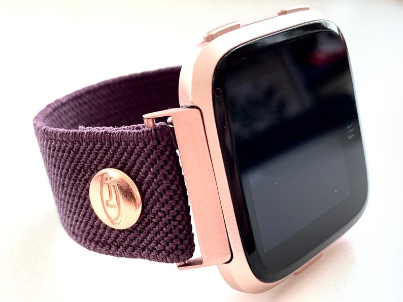 May include: A rose gold smartwatch with a dark gray screen and a dark purple elastic band. The band has a gold button with a logo on it.
