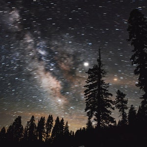 May include: A long exposure photograph of a night sky with stars and the Milky Way visible. The sky is dark blue and the stars are white. There are silhouetted trees in the foreground.
