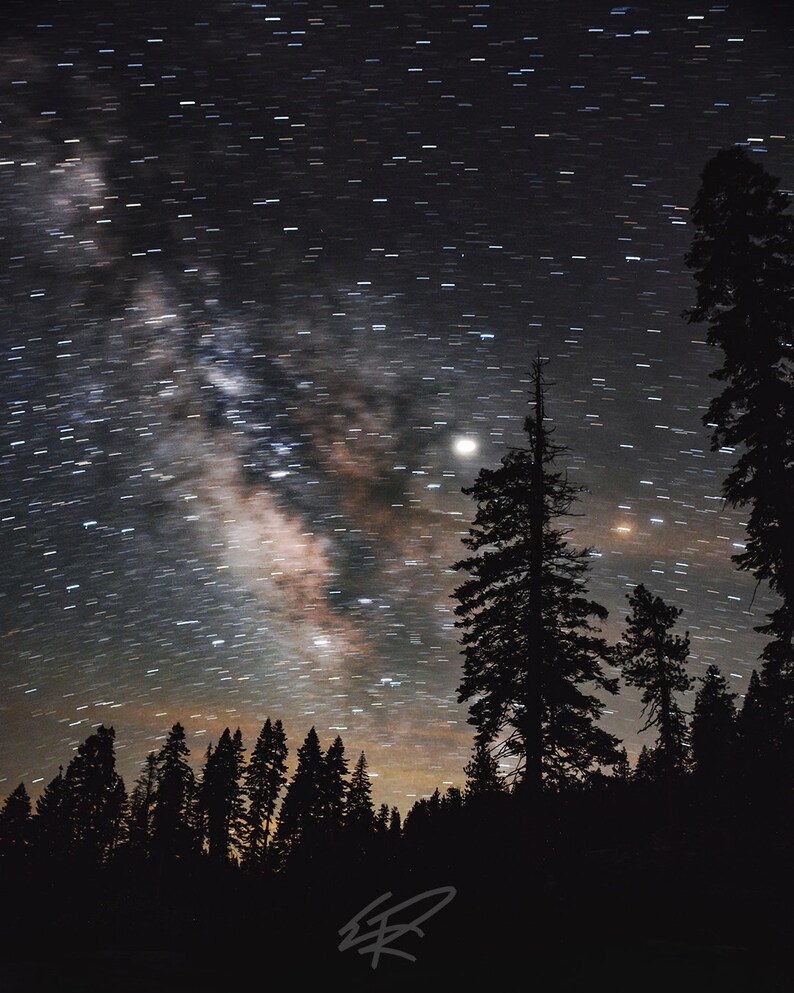 May include: A long exposure photograph of a night sky with stars and the Milky Way visible. The sky is dark blue and the stars are white. There are silhouetted trees in the foreground.