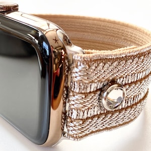 May include: A rose gold Apple Watch with a beige elastic band featuring a silver and beige woven pattern and a clear crystal accent.