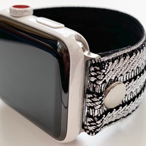 Puede incluir: Un Apple Watch blanco con una banda de patr&oacute;n negro y plateado. La banda tiene un cierre de metal plateado.