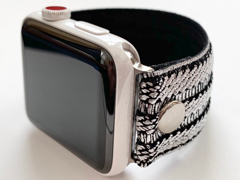 Puede incluir: Un Apple Watch blanco con una banda de patr&oacute;n negro y plateado. La banda tiene un cierre de metal plateado.