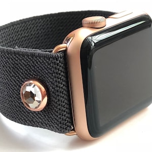 May include: A rose gold Apple Watch with a black fabric band and a clear crystal embellishment.