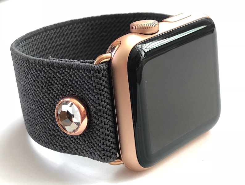 May include: A rose gold Apple Watch with a black fabric band and a clear crystal embellishment.