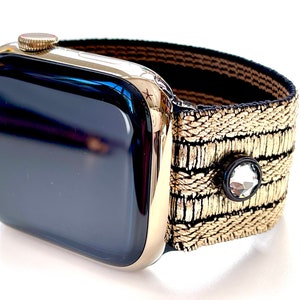 May include: A gold Apple Watch with a black face and a gold and black patterned band. The band has a small clear rhinestone embellishment.
