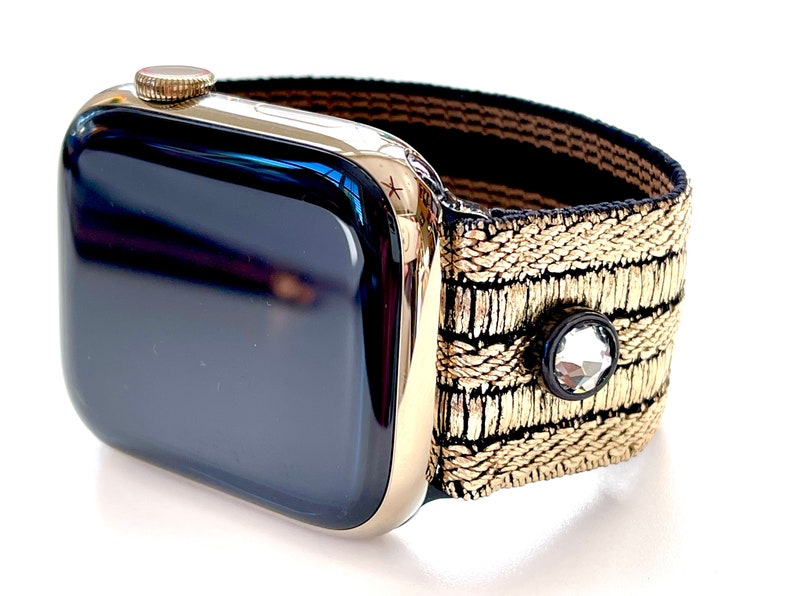 May include: A gold Apple Watch with a black face and a gold and black patterned band. The band has a small clear rhinestone embellishment.