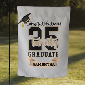 May include: White graduation yard sign with a black graduation cap, gold text that says "Congratulations 25 twenty GRADUATE SAMANTHA" and a gold ribbon.