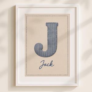 May include: A framed wall art print featuring a large, dark blue letter J with thin white vertical stripes centered on a light beige background. Below the letter, the name Jack is written in a dark blue cursive script. The print has a stitched border.