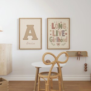 Puede incluir: Dos l&aacute;minas decorativas rectangulares en marcos de madera clara. La l&aacute;mina de la izquierda muestra una letra A grande con el nombre Amelia debajo. La de la derecha presenta el texto LONG LIVE Girlhood con motivos florales, corazones y mariposas.