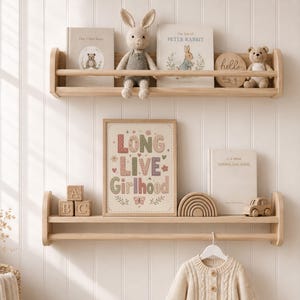 Puede incluir: Dos estantes de pared de madera clara con una barra frontal. El estante superior sostiene un conejo de peluche, dos libros y un letrero redondo. El estante inferior muestra bloques de madera, un cuadro con el texto LONG LIVE Girlhood y un libro.