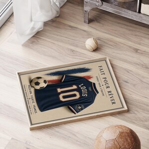 May include: A framed rectangular poster featuring a dark blue soccer jersey with the number 10 and the name Louis printed on the back. The design includes a soccer ball, red and blue brush strokes, and the text Fait pour r&ecirc;ver, Passion, Fiert&eacute;, France.
