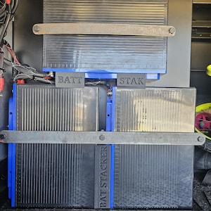 Puede incluir: Primer plano de un sistema de apilamiento de baterías negro y azul. El sistema se compone de múltiples unidades rectangulares con una superficie acanalada, aseguradas por barras metálicas. El texto "BATT STAK" y "BAT STACKER" es visible.