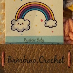 May include: A brown leather tag with the text "Bambino Crochet" in white lettering. The tag is attached to a white card with a rainbow and clouds illustration and the text "Rainbow Baby".