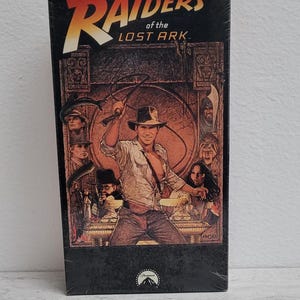 May include: A VHS tape case for the movie Raiders of the Lost Ark. The cover features a black and white illustration of Indiana Jones holding a whip, surrounded by other characters from the film.