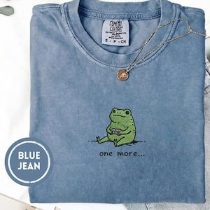May include: Blue jean Comfort Colors t-shirt with a green frog graphic holding a game controller. The text "one more..." is below the frog. A gold necklace with a pendant is on the shirt.