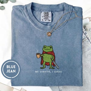 May include: Blue jean-colored t-shirt with a graphic of a green frog wearing a red cape and holding a coffee cup and sword. The text "WE SURVIVE, I GUESS" is below the frog. A gold necklace is draped across the shirt.