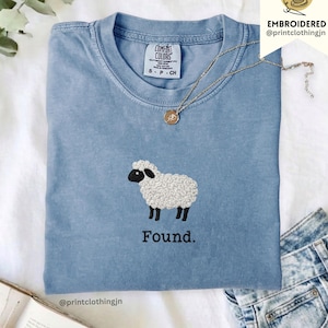 Peut inclure: Un t-shirt bleu à col rond avec une broderie représentant un mouton blanc à tête et pattes noires. Sous le mouton, le mot Found est brodé en fil noir. Le t-shirt porte une étiquette Comfort Colors au col et un collier doré.