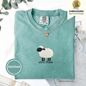 Könnte beinhalten: Meergrünes T-Shirt mit gesticktem Schaf-Design und dem Text "still found." Das Shirt hat einen Rundhalsausschnitt und kurze Ärmel. Eine silberne Halskette mit Anhänger liegt auf dem Shirt. Die Marke "Comfort Colors" ist sichtbar.