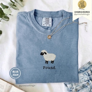 Könnte beinhalten: Ein blaues T-Shirt mit kurzen Ärmeln und einer Stickerei eines weißen Schafs mit schwarzem Gesicht und Beinen. Unter dem Schaf ist das Wort Found in schwarzem Garn gestickt. Das T-Shirt hat einen Rundhalsausschnitt und ein Comfort Colors Etikett.