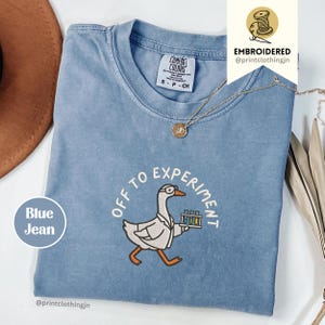 Könnte beinhalten: Ein blaues T-Shirt mit einer Stickerei, die eine weiße Gans mit Brille und Laborkittel zeigt, die ein Tablett mit Reagenzgläsern hält. Der Text "OFF TO EXPERIMENT" ist bogenförmig über der Gans angebracht. Das Shirt hat ein Comfort Colors Etikett.