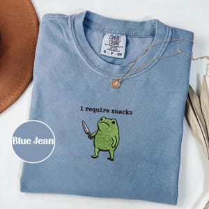 May include: Blue jean colored t-shirt with the text "i require snacks" and an embroidered green frog holding a knife. A gold necklace with a pendant is draped across the shirt. The shirt has a Comfort Colors tag.