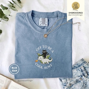 Könnte beinhalten: Ein blaues T-Shirt mit Rundhalsausschnitt und einer Stickerei, die eine schwarze Katze mit Brille auf einem weißen Drachen mit grünen Flügeln zeigt. Der Text "OFF TO BE A COOL AUNT" ist in Weiß um die zentrale Illustration gestickt. Das T-Shirt hat ein Comfort Colors-Etikett am inneren Halsausschnitt.