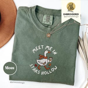 Könnte beinhalten: Ein moosgrünes T-Shirt mit Rundhalsausschnitt und einer Stickerei, die eine weiße Gans mit orangefarbenem Schal und einer Kaffeetasse zeigt. Der Text "MEET ME IN STARS HOLLOW" ist in Weiß um die Gans herum gestickt, ergänzt durch kleine orange und rote Blätter.