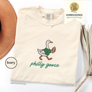 Può includere: Una t-shirt color avorio a girocollo con il ricamo di un'oca che indossa una maglia verde e tiene in mano un pallone da football americano marrone. Sotto l'oca, la scritta "philly goose" è ricamata in corsivo verde. Etichetta Comfort Colors.