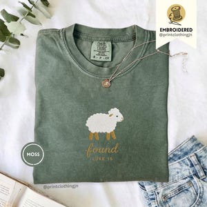 Könnte beinhalten: Ein moosgrünes Kurzarm-T-Shirt mit einem gestickten weißen Schaf mit braunen Beinen. Unter dem Schaf steht der Text "found" in Schreibschrift und "LUKE 15" in einer einfachen Schriftart. Das T-Shirt hat einen Rundhalsausschnitt und ein Comfort Colors Etikett.