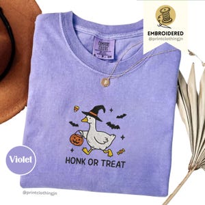 May include: A violet short-sleeved t-shirt featuring an embroidered graphic of a white goose wearing a black witch hat, holding a jack-o'-lantern bucket. The text reads HONK OR TREAT, surrounded by small bats, candy corn, and stars.