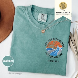 Könnte beinhalten: Ein kurzärmeliges T-Shirt in Meerschaumgrün mit einer Stickerei auf der Brust. Das Motiv zeigt eine Sonne, die über stilisierten blauen und grauen Wellen aufgeht, mit dem Text "THROUGH DEEP WATERS I WILL BE WITH YOU" und "ISAIAH 43:2".