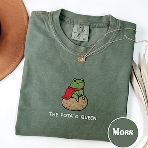 May include: A moss green t-shirt featuring an embroidered frog in a red cape, seated on a potato, with the text "THE POTATO QUEEN". A gold necklace and a brown hat are also visible.