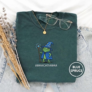 Puede incluir: Sudadera de color verde azulado con un bordado de una rana mago sosteniendo un bastón. La palabra "ABRACADABRA" está impresa debajo. Un collar dorado y gafas transparentes descansan sobre la camisa. La camisa tiene una etiqueta "Blue Spruce".
