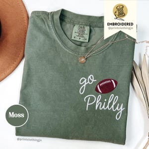 Könnte beinhalten: Ein moosgrünes Kurzarm-T-Shirt mit Rundhalsausschnitt. Das Shirt ist mit weißem Sticktext "go Philly" und einer Grafik eines bordeaux-weißen Footballs versehen. Eine goldfarbene Halskette mit einem kleinen runden Anhänger liegt auf dem Kragen.