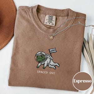 May include: Espresso-colored t-shirt with a crew neck. The shirt features an embroidered astronaut frog with a "Mars" flag and the text "SPACED OUT". A silver necklace with a pendant is also visible.
