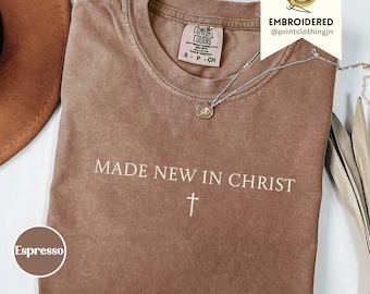May include: An espresso-coloured short-sleeve t-shirt featuring the text MADE NEW IN CHRIST in white lettering above a small white cross symbol. The shirt has a Comfort Colors brand label at the inner neckline and a silver-toned chain necklace.