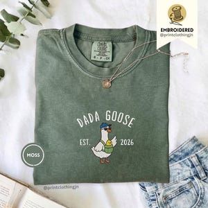 May include: A moss green crew neck t-shirt featuring an embroidered graphic of a goose wearing a blue baseball cap and sunglasses, carrying a small yellow chick in a green pouch. The text reads DADA GOOSE EST. 2026 in white lettering.