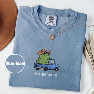 Könnte beinhalten: Blaues Jeans-T-Shirt mit einem gestickten Design einer grünen Frosch mit Cowboyhut, der einen blauen Truck fährt. Der Text "NO REGRETS" steht darunter. Eine goldene Halskette liegt auf dem Shirt.